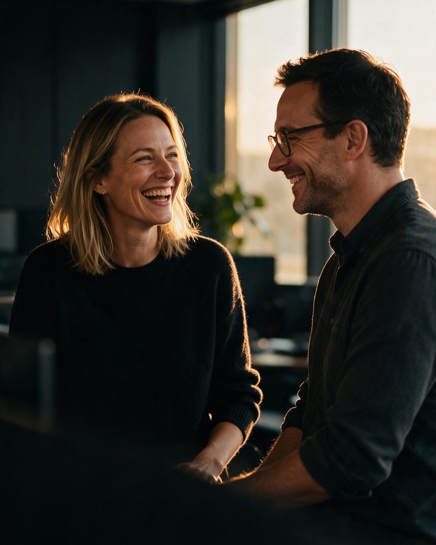Zwei Kolleg:innen lachen miteinander im warmen Abendlicht eines Büros (KI-generiert)